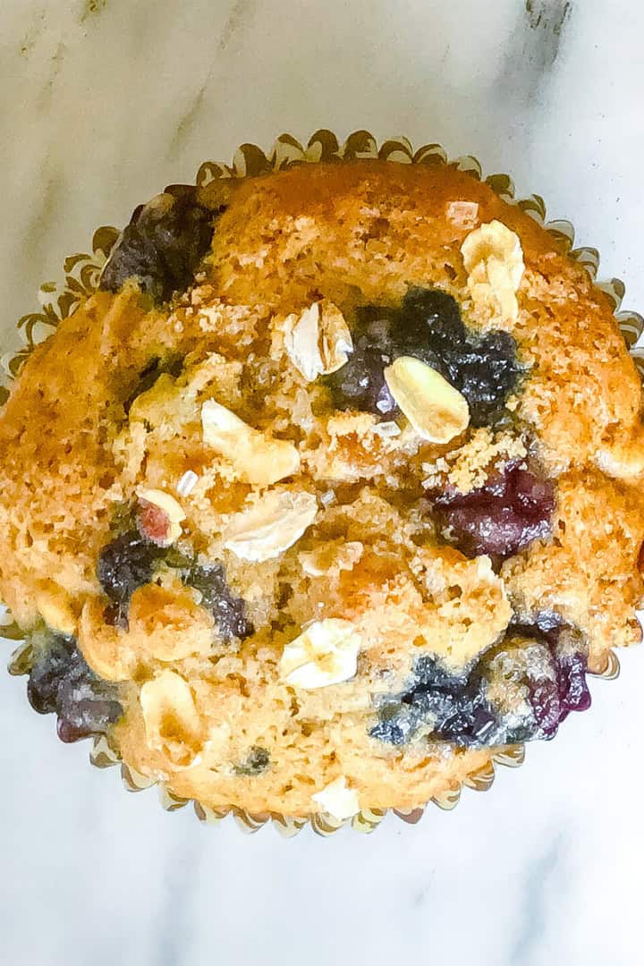 A single blueberry oat muffin with oats on top, in a paper liner, sits on a white marble surface.