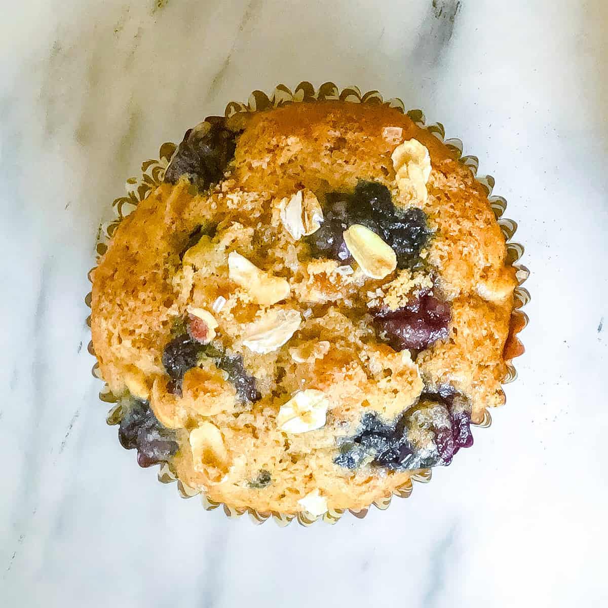 A single blueberry oat muffin with oats on top, in a paper liner, sits on a white marble surface.