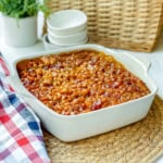 A white baking dish filled with Boston baked beans sits on a woven placemat, next to a checkered cloth and stacked white bowls.