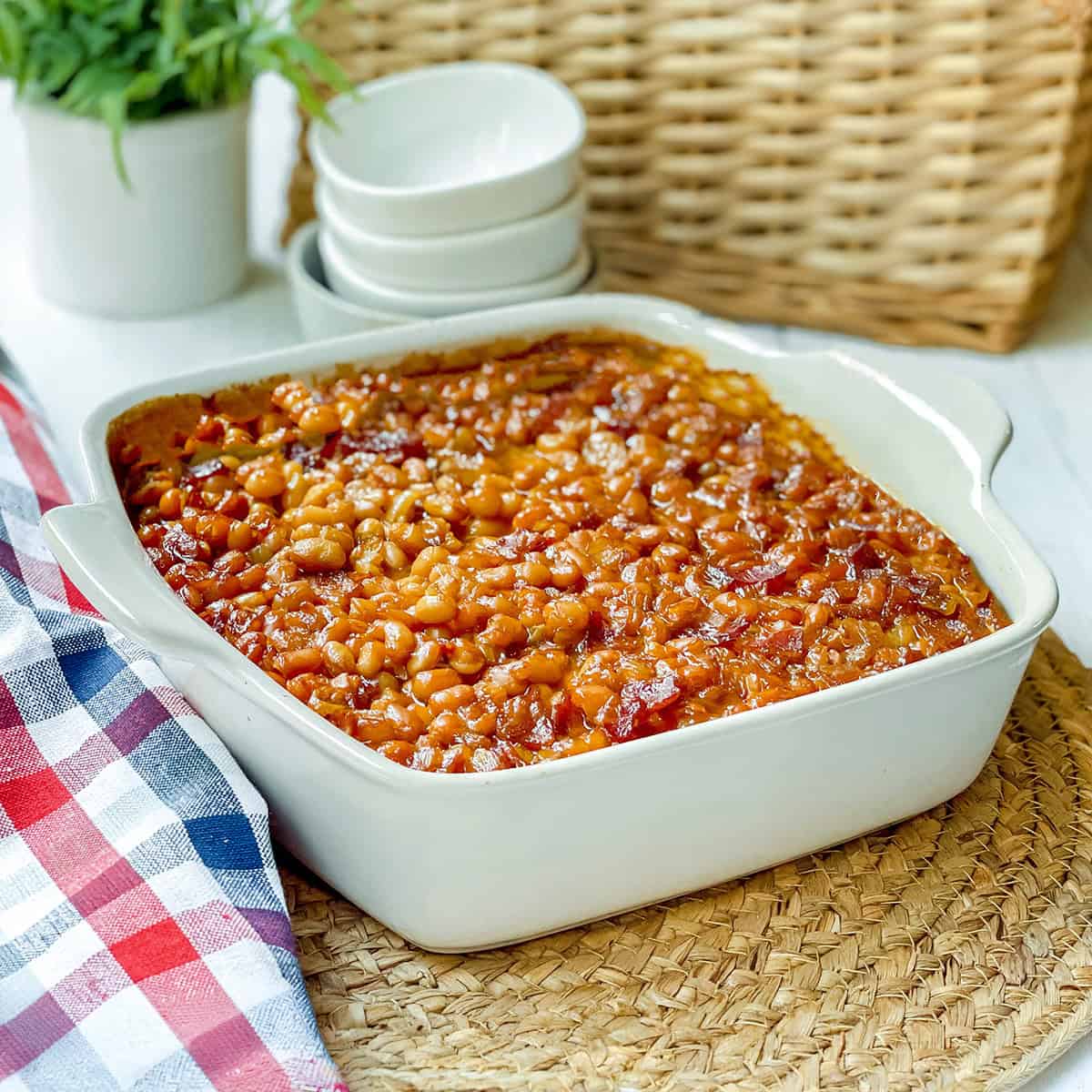 A white baking dish filled with Boston baked beans sits on a woven placemat, next to a checkered cloth and stacked white bowls.