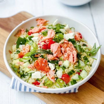 A bowl of vibrant couscous salad topped with flaked salmon, cucumber, tomato, feta cheese, green onions, and arugula sits beautifully on a wooden board.