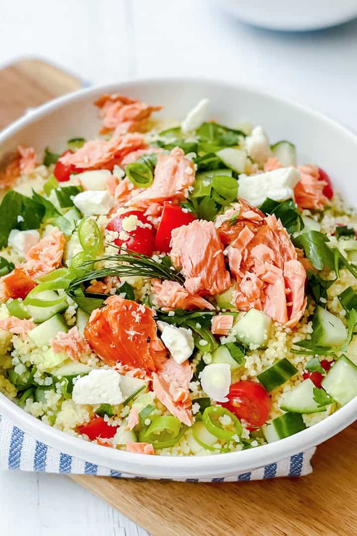A bowl of vibrant couscous salad topped with flaked salmon, cucumber, tomato, feta cheese, green onions, and arugula sits beautifully on a wooden board.