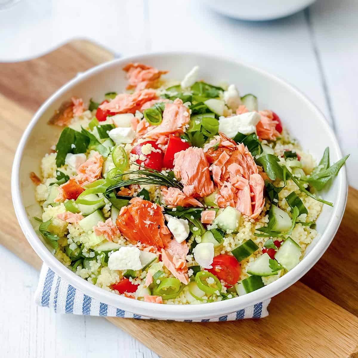 A bowl of vibrant couscous salad topped with flaked salmon, cucumber, tomato, feta cheese, green onions, and arugula sits beautifully on a wooden board.