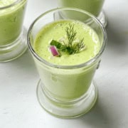 A glass of cucumber soup garnished with fresh dill, a slice of cucumber, and a small piece of red onion, set on a light surface.