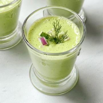 A glass of cucumber soup garnished with fresh dill, a slice of cucumber, and a small piece of red onion, set on a light surface.