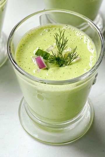 A glass of cucumber soup garnished with fresh dill, a slice of cucumber, and a small piece of red onion, set on a light surface.