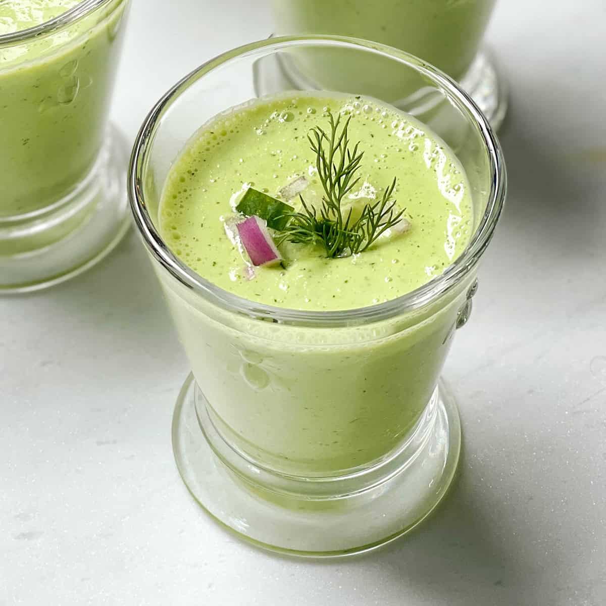A glass of cucumber soup garnished with fresh dill, a slice of cucumber, and a small piece of red onion, set on a light surface.