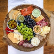 A round wooden platter serves as a Mediterranean charcuterie board, featuring assorted cheeses, crackers, bread, grapes, olives, cured meats, nuts, and spreads.