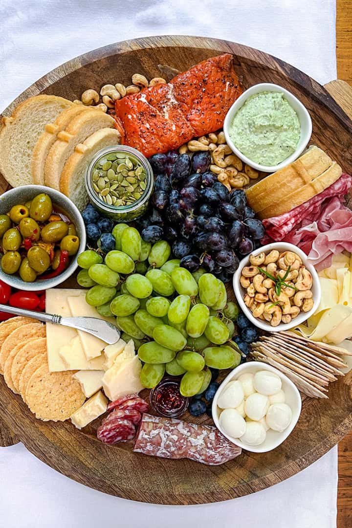 A round wooden platter serves as a Mediterranean charcuterie board, featuring assorted cheeses, crackers, bread, grapes, olives, cured meats, nuts, and spreads.