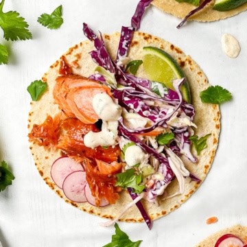 Tortilla topped with shredded salmon, sliced radishes, red cabbage slaw, cilantro, lime wedge, and creamy dressing on a white background.