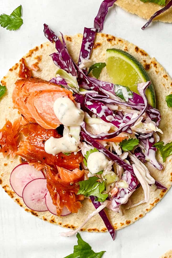 Tortilla topped with shredded salmon, sliced radishes, red cabbage slaw, cilantro, lime wedge, and creamy dressing on a white background.