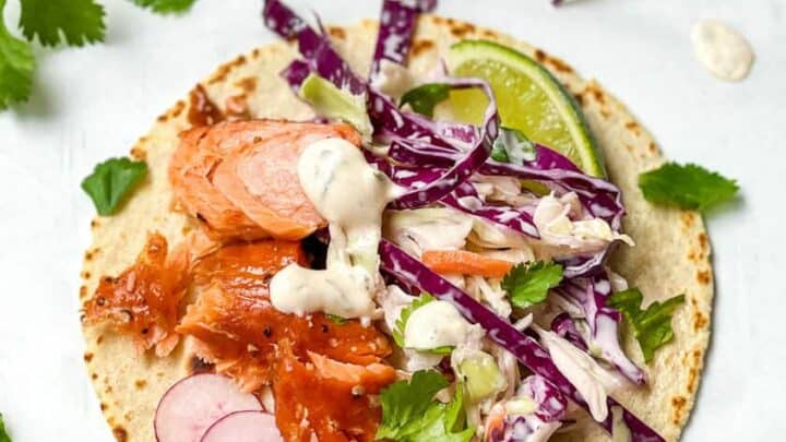 A corn tortilla transforms into delicious salmon tacos, topped with flaked salmon, sliced radishes, shredded red cabbage, cilantro, a lime wedge, and a drizzle of creamy sauce.