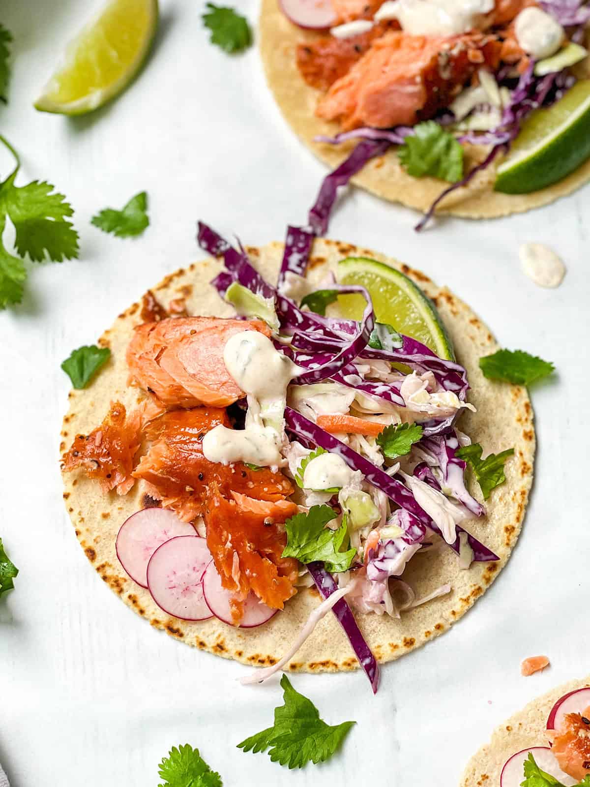 A taco topped with cooked salmon, sliced radishes, shredded cabbage, creamy sauce, and a lime wedge on a tortilla; cilantro leaves are scattered around-these fresh salmon tacos burst with vibrant flavors and color in every bite.
