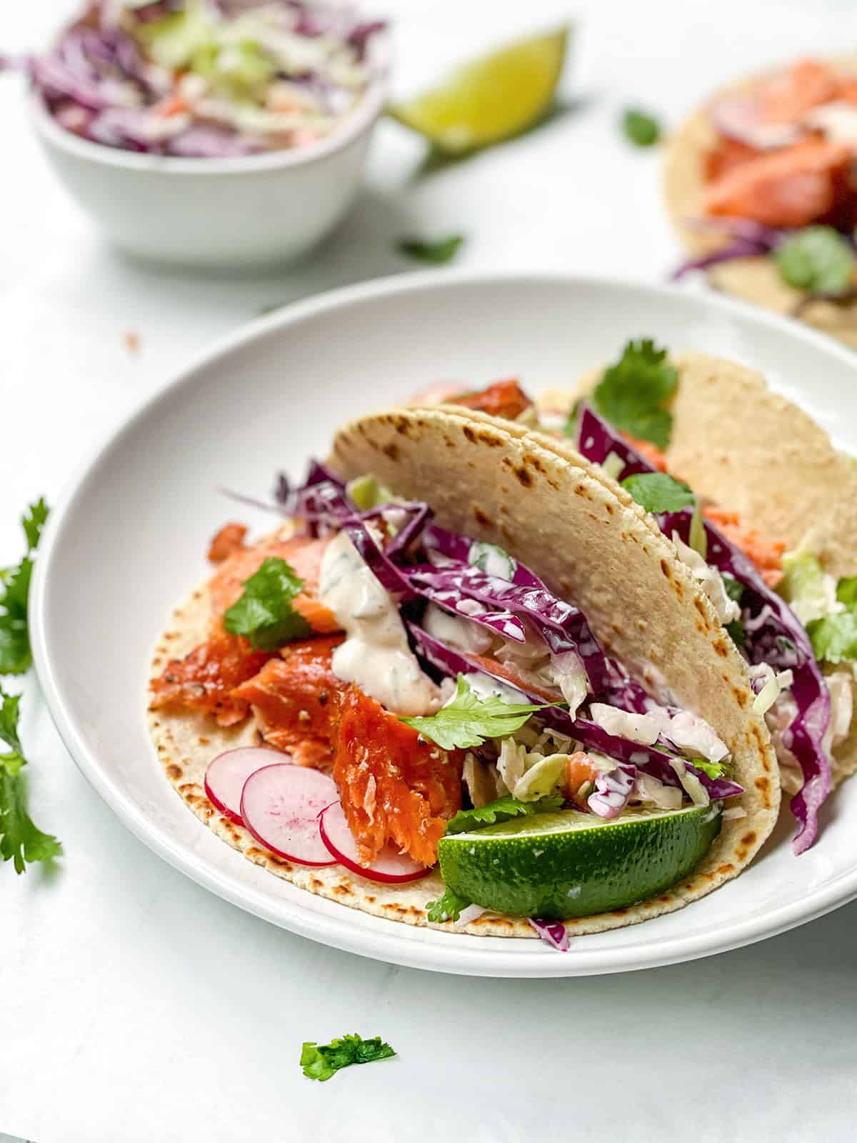 Two salmon tacos filled with smoked salmon, purple cabbage, radish slices, lettuce, and sauce, garnished with lime wedges and cilantro on a white plate.