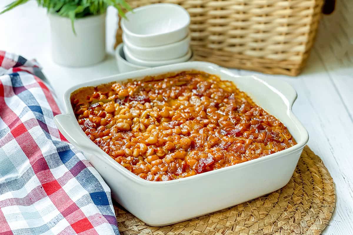 A square white casserole dish filled with Boston baked beans sits on a woven mat, with stacked bowls and a red, white, and blue checkered cloth nearby.