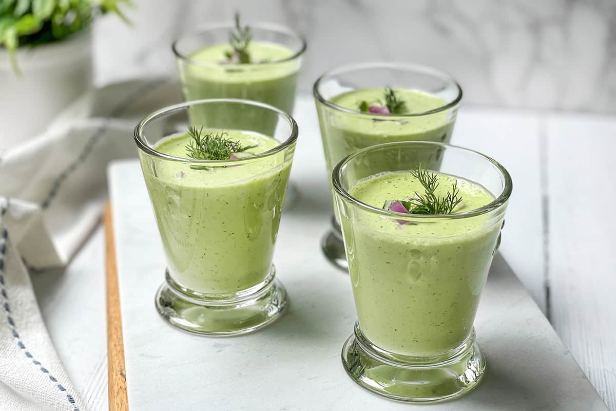 Four glasses of green cucumber soup garnished with dill and small pieces of red onion, arranged on a white surface.