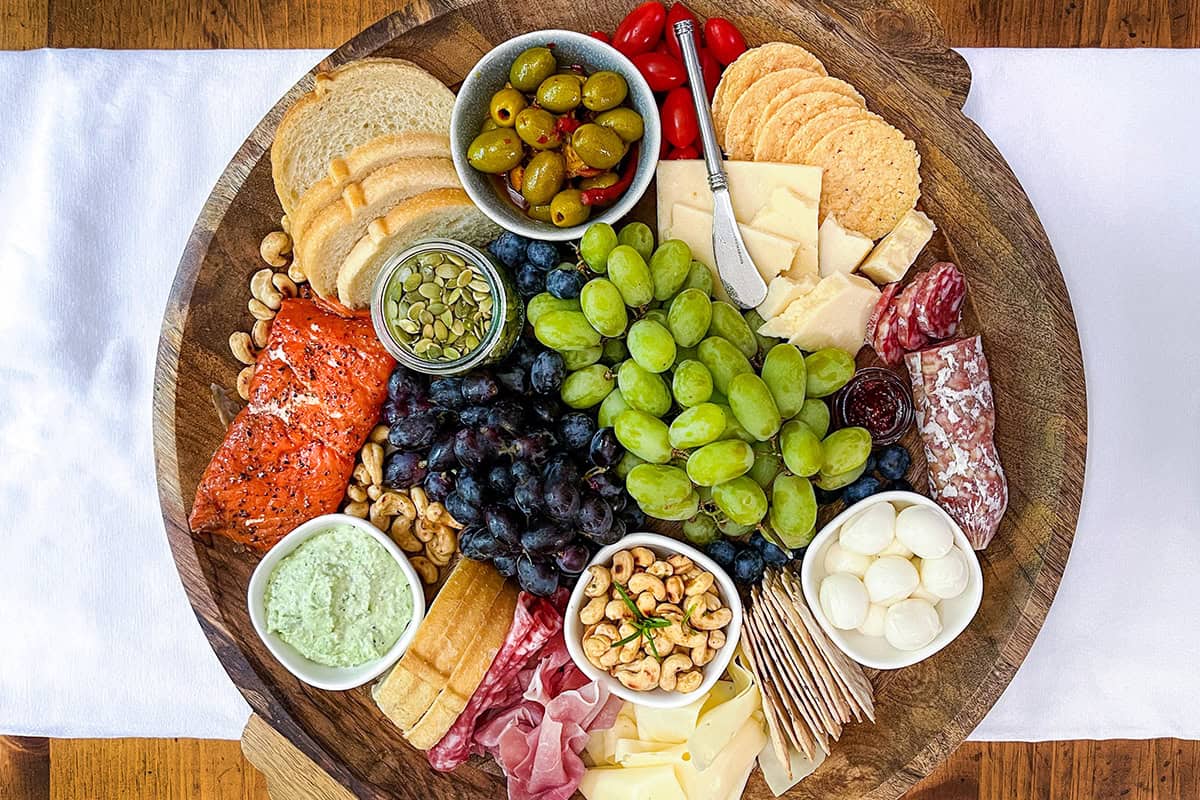 A round wooden platter showcases a Mediterranean charcuterie board with assorted cheeses, crackers, salami, grapes, olives, bread, nuts, smoked salmon, tomatoes, dip, seeds-all arranged neatly around a cheese knife.