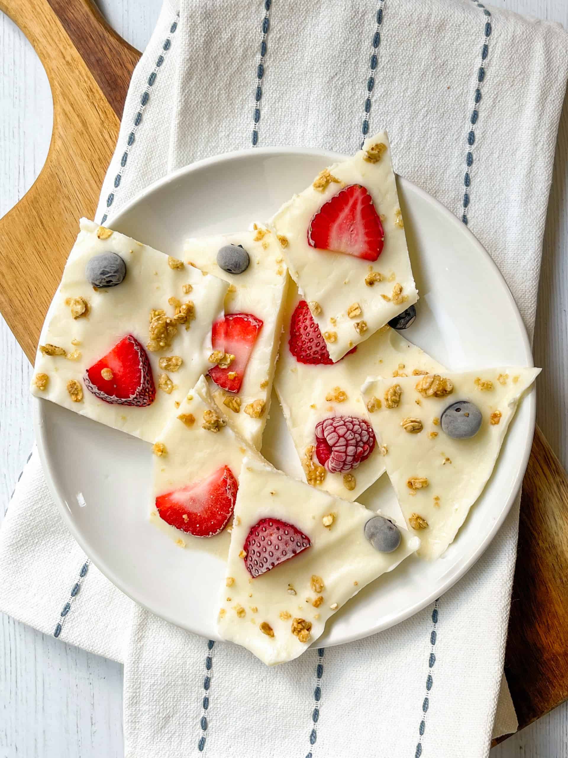Frozen yogurt bark pieces topped with strawberries, blueberries, and granola, served on a white plate over a striped cloth on a wooden surface.