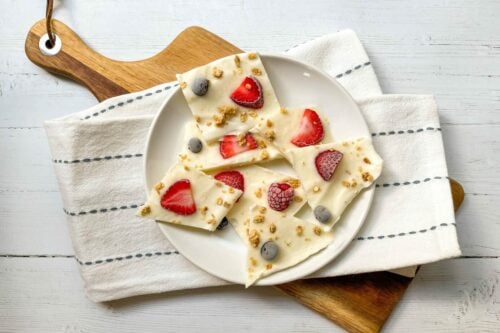 A white plate with pieces of frozen yogurt bark topped with strawberries, blueberries, and granola, set on a napkin and wooden boards.
