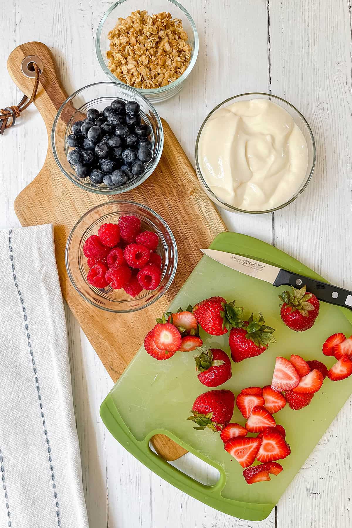 Bowls of blueberries, raspberries, granola, and yogurt are arranged next to sliced strawberries on a cutting board, perfect ingredients for making frozen yogurt bark, with a knife and kitchen towel nearby on a white wooden surface.