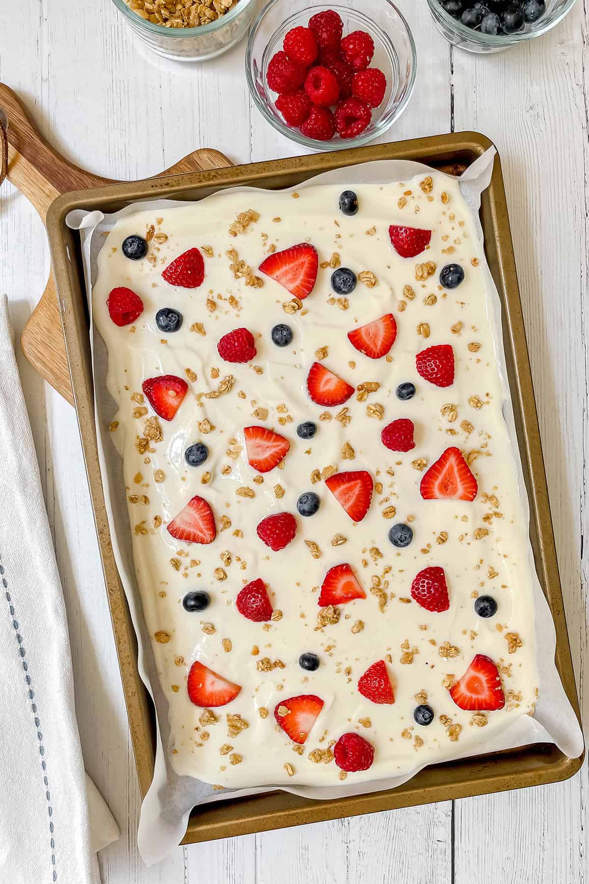 A baking tray lined with parchment paper holds a creamy frozen yogurt bark topped with strawberries, raspberries, blueberries, and scattered granola clusters. Bowls of fruit are in the background.