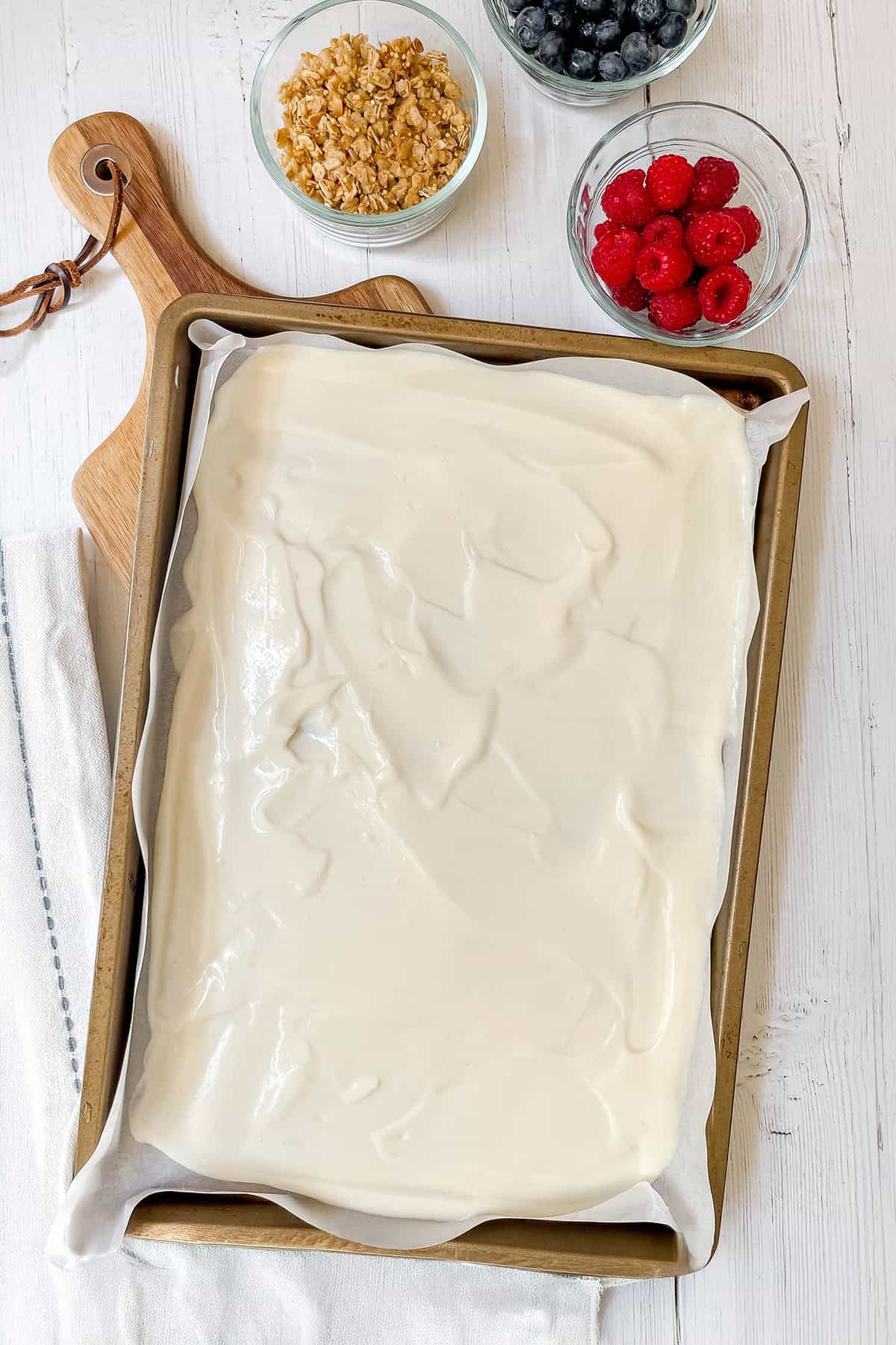 A baking sheet covered with a layer of yogurt for frozen yogurt bark, surrounded by small bowls of granola, blueberries, and raspberries on a white surface.