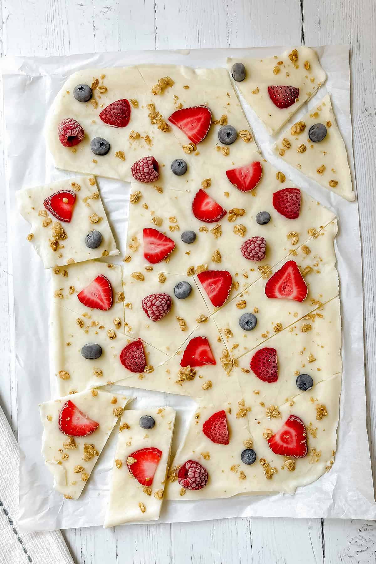 Rectangular pieces of frozen yogurt bark topped with strawberries, blueberries, raspberries, and granola are arranged on parchment paper on a white wooden surface.