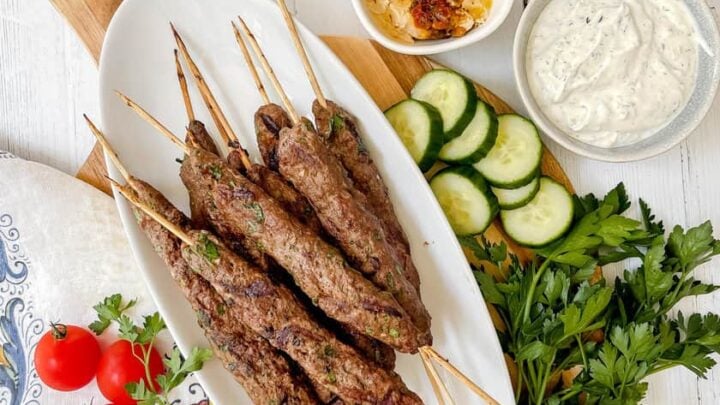 Top view of Beef Kofta Kebobs on white platter with hummus and tzatziki sauce