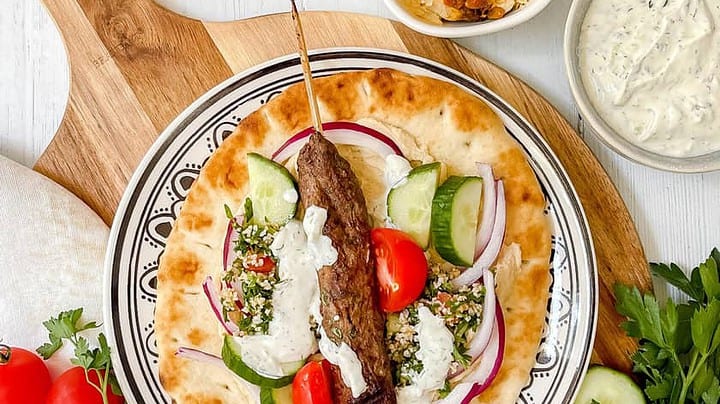 Top view of Beef Kofta Kabob on warm pita with tzatziki sauce, hummus, tomatoes and cucumbers on a plate