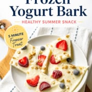 A plate of frozen yogurt bark topped with strawberries, blueberries, and granola sits on a striped cloth, with text highlighting this frozen yogurt bark as a 3-ingredient, 5-minute healthy summer snack.