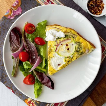 A slice of zucchini frittata with dollops of ricotta, is served alongside a fresh side salad of mixed greens, tomato, and onion on a white plate.