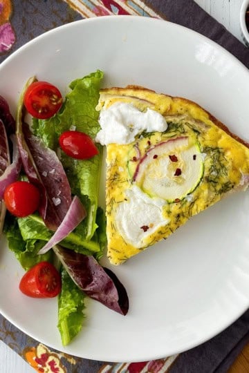 A slice of zucchini frittata with dollops of ricotta, is served alongside a fresh side salad of mixed greens, tomato, and onion on a white plate.