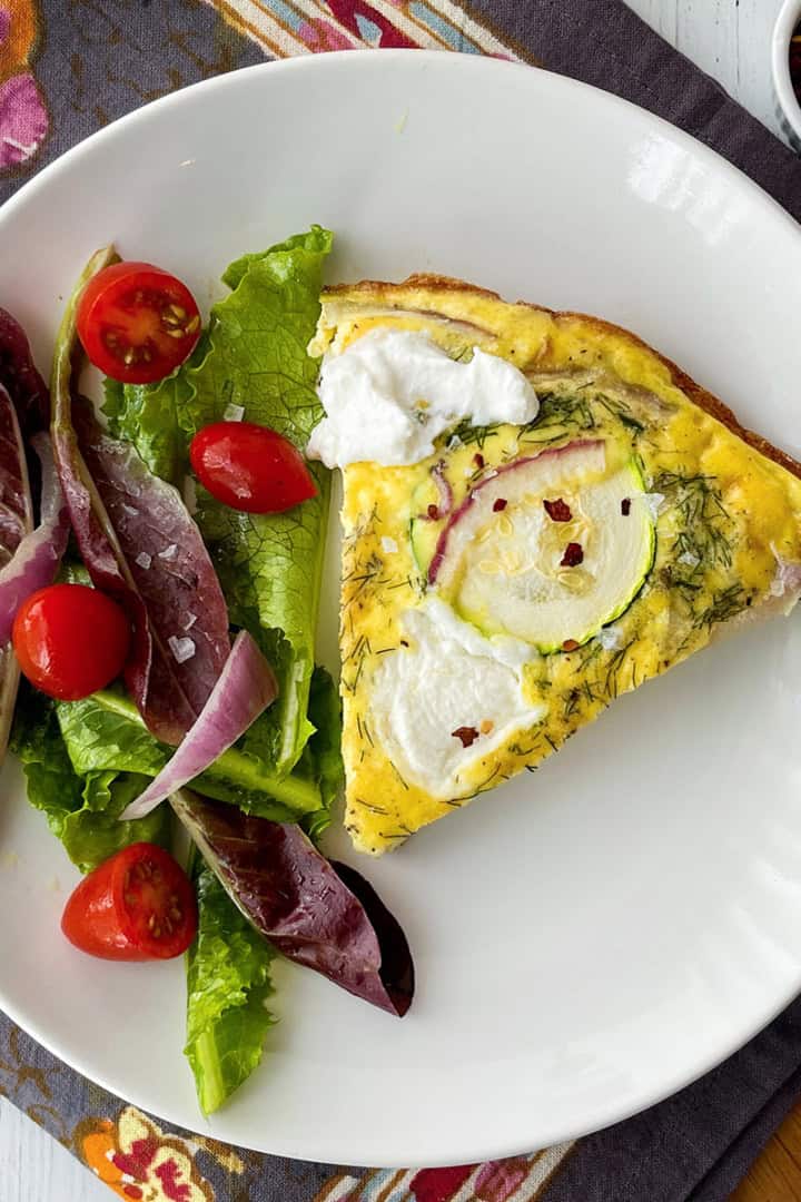 A slice of zucchini frittata with dollops of ricotta, is served alongside a fresh side salad of mixed greens, tomato, and onion on a white plate.