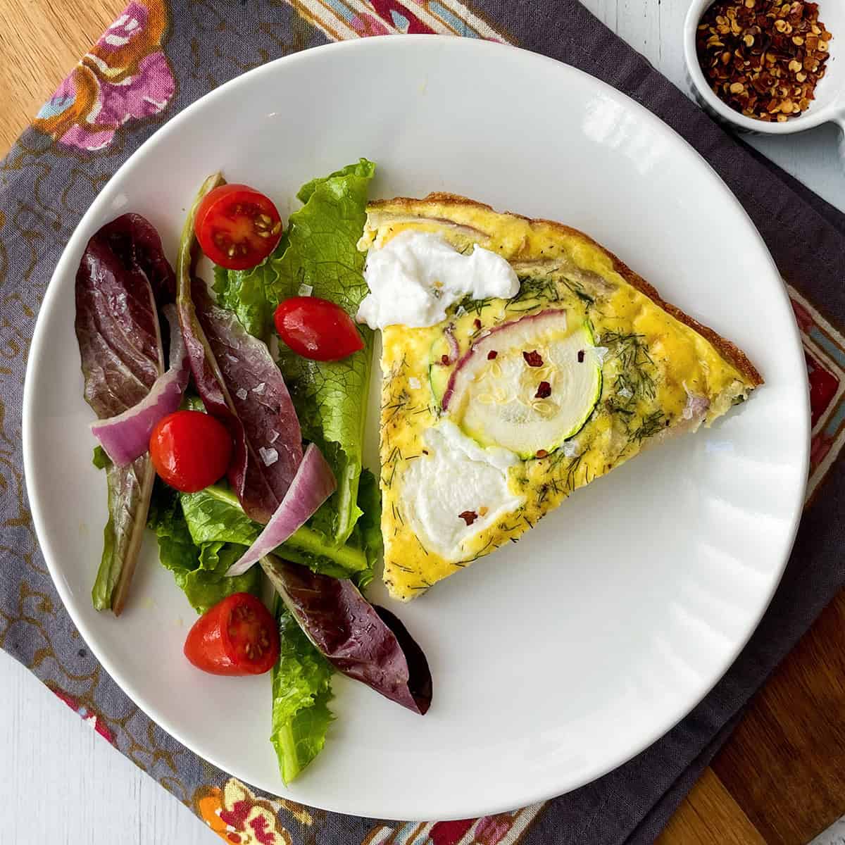 A slice of zucchini frittata with dollops of ricotta, is served alongside a fresh side salad of mixed greens, tomato, and onion on a white plate.