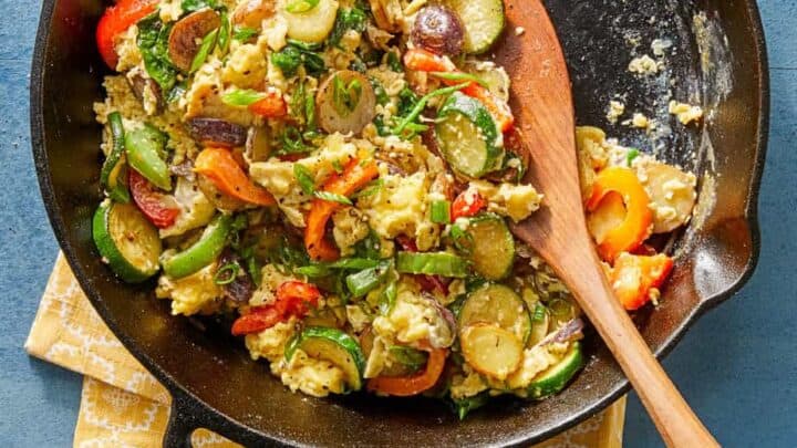 A cast iron skillet filled with scrambled eggs, sliced vegetables, and herbs sits on a yellow cloth with a wooden spoon resting inside—perfect for easy One Pot Summer Dinners.