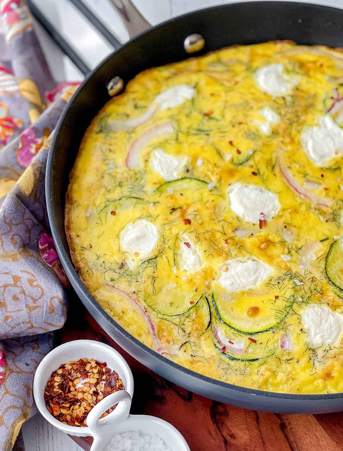 Zucchini frittata in a black skillet with visible zucchini, red onion, herbs, and dollops of ricotta.