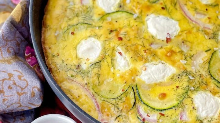 A baked zucchini frittata in a skillet with visible zucchini slices, red onions, and dollops of cheese, served next to bowls of red pepper flakes and coarse salt—perfect for any zucchini frittata recipe lover.