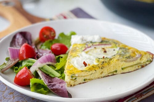 Side view of Zucchini Frittata wedge with a green salad on a white plate.