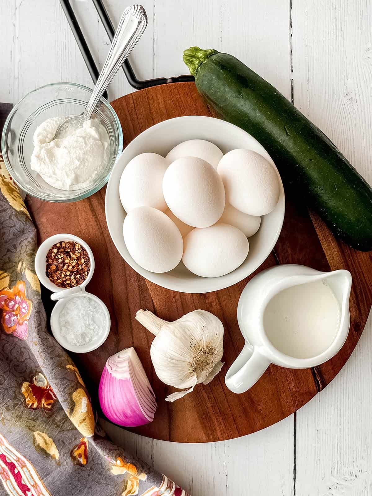 A bowl of eggs, a zucchini, a glass of ricotta, cream in a pitcher, red onion, garlic, salt, and red pepper flakes are arranged on a wooden board-everything you need for a delicious zucchini frittata recipe.