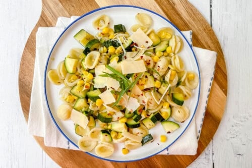 A plate of zucchini pasta featuring orecchiette, corn, parmesan shavings, red pepper flakes, and a fresh herb sprig, served on a white plate with a napkin and wooden board.