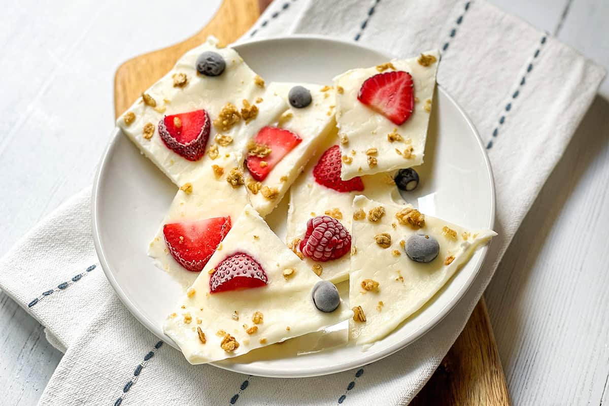 A white plate with pieces of frozen yogurt bark topped with strawberries, raspberries, blueberries, and granola, placed on a folded napkin and wooden board.