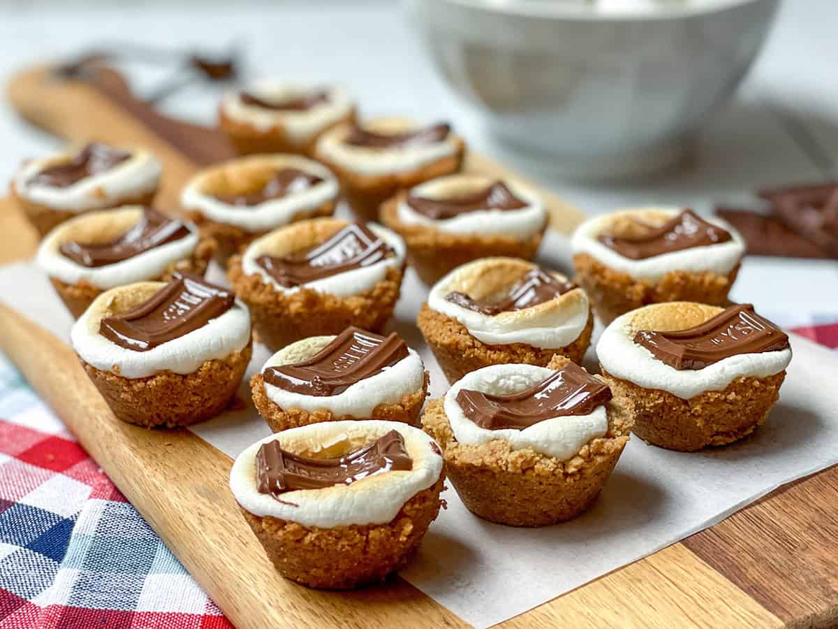 A wooden board displays s'mores cookie cups topped with toasted marshmallows and chocolate pieces, with a bowl and checkered cloth in the background.