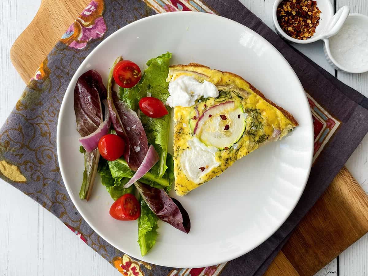 A plate with a slice of zucchini frittata and a side salad featuring lettuce, cherry tomatoes, and red onion rests on a patterned napkin.