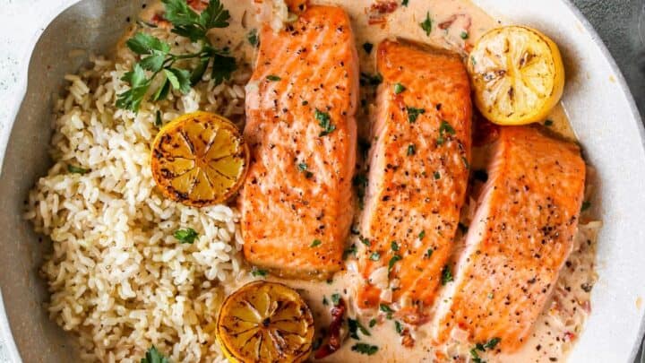 Three cooked salmon fillets with creamy sauce, brown rice, parsley, and two seared lemon halves in a skillet—an easy one skillet Mediterranean Diet dinner—served with plates and utensils nearby.