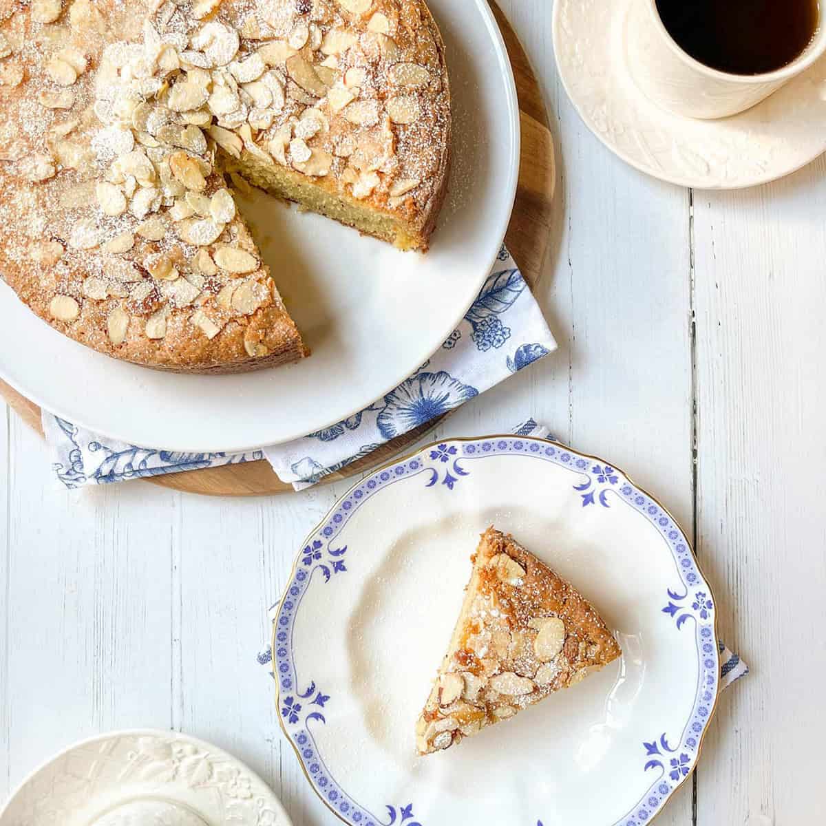A round almond Anytime Snack Cake with a slice removed sits on a wooden board; the slice is served on a plate next to a cup of coffee on a white table.