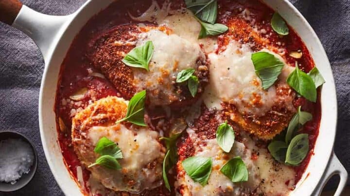 A skillet with breaded chicken breasts topped with melted cheese and fresh basil, served over tomato sauce on a gray cloth napkin—an easy one skillet Mediterranean Diet dinner.