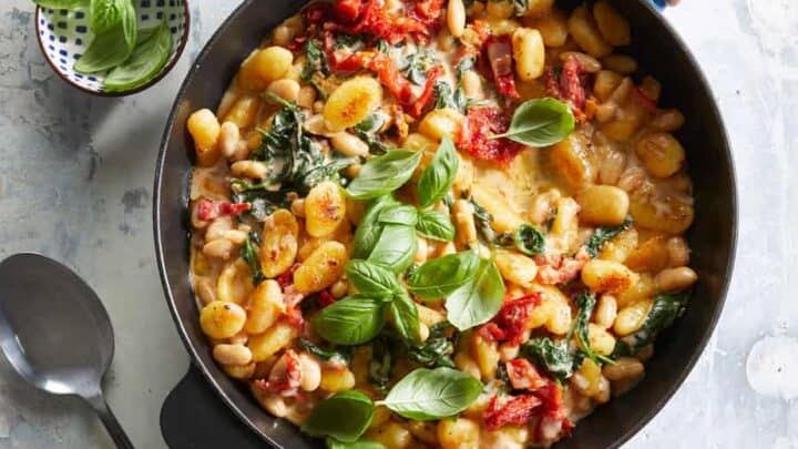A skillet filled with gnocchi, white beans, spinach, sun-dried tomatoes, and fresh basil—this vibrant dish is perfect for anti-inflammatory dinners. A spoon and a small bowl of basil leaves sit invitingly beside it.