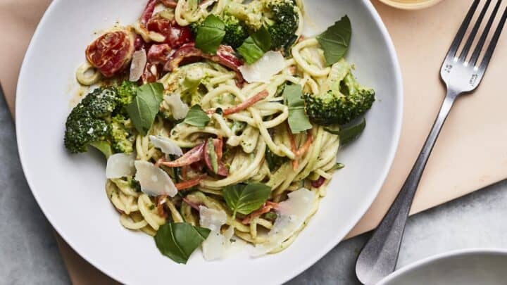 A plate of vegetable pasta with broccoli, cherry tomatoes, basil leaves, and Parmesan cheese shavings sits next to a fork and a glass of light-colored beverage—perfect for healthy 20 minute dinners.