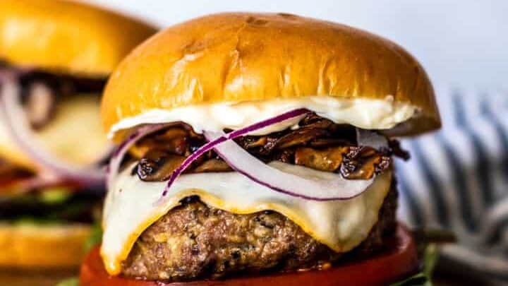 A cheeseburger on a wooden board, topped with melted cheese, grilled onions, lettuce, and tomato—perfect for your Labor Day recipes—with another burger in the background.