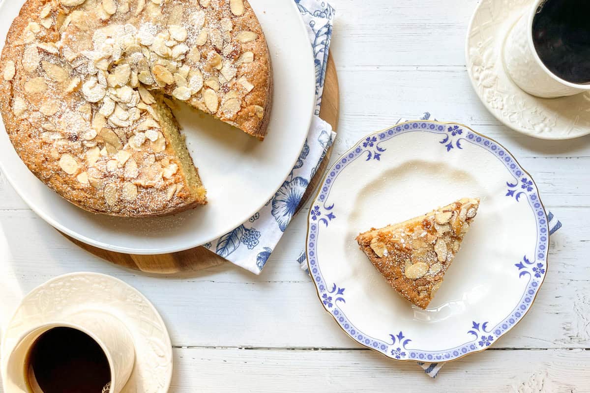 A round almond cake-one slice cut out-sits on a plate; the slice, perfect as an Anytime Snack Cake, is served on a smaller plate next to two cups of black coffee on a white table.
